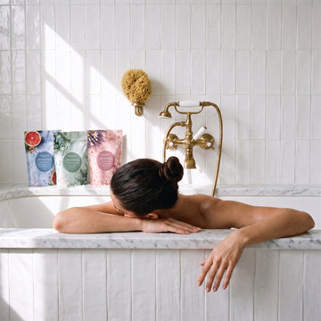A person with a hair bun rests their arms on a marble bathtub edge next to three Odacite Relax & Recenter Bath Set pouches.
