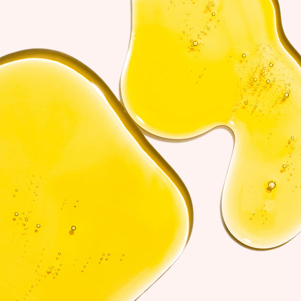 Odacité C-Glow Hydra-Firm Body Oil—Vitamin C & E + Kakadu—with yellow liquid puddles and bubbles on a neutral background.