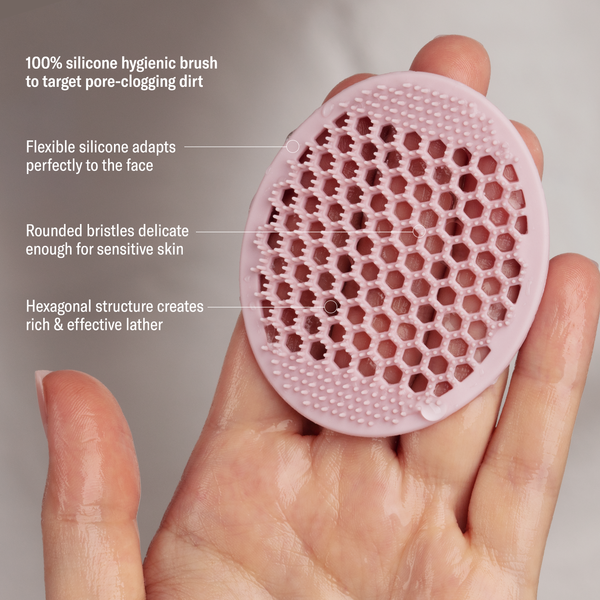 A hand holds the Odacité Pore & Face Cleansing Brush, a pink silicone brush with textured bristles.