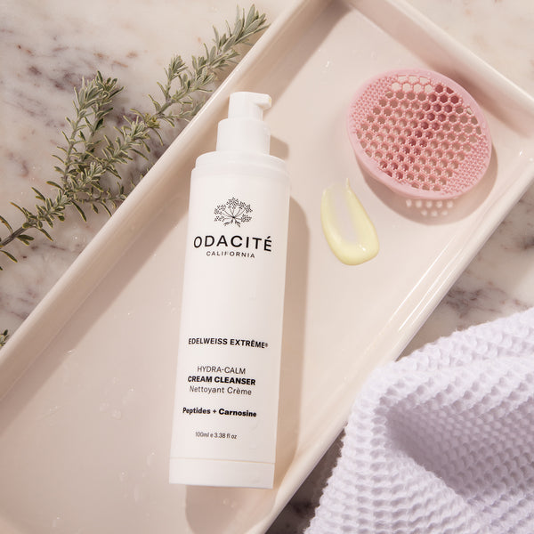 Odacité Daily Calm Cleansing Duo on a tray with a silicone brush, green leaves, cleanser dollop, and face towel.