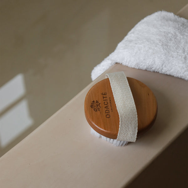 Odacité Dry Spa Brush with soft bristles on a folded white towel next to a bathtub, designed for sensitive skin.