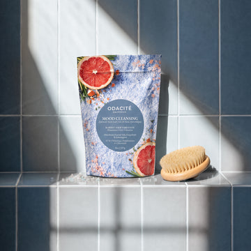 Odacite Mood Cleansing Himalayan Crystal Salt, Grapefruit & Lemongrass Ayurvedic Bath Soak pouch and a wooden bath brush on a tiled ledge.