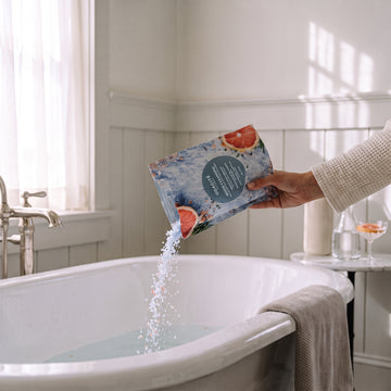 Hand pouring blue Odacite Mood Cleansing Ayurvedic Bath Soak into a white clawfoot tub.