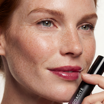 Woman with clear skin holds Odacité SPF 40 Nourishing Lip Serum by her face, with natural makeup and smooth, radiant lips.