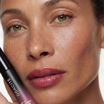 A woman with glowing, freckled skin holds Odacité SPF 40 Nourishing Lip Serum in a small tube, her face softly illuminated.