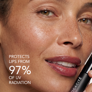 Woman with shiny lips holds Odacité SPF 40 Nourishing Lip Serum in clear tube, cap off, close to her mouth.