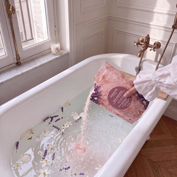 A hand pours Soul Soothing • Himalayan Crystal Salt, & French Lavender Neroli + Patchouli • Ayurvedic Bath Soak into a petal-filled tub.