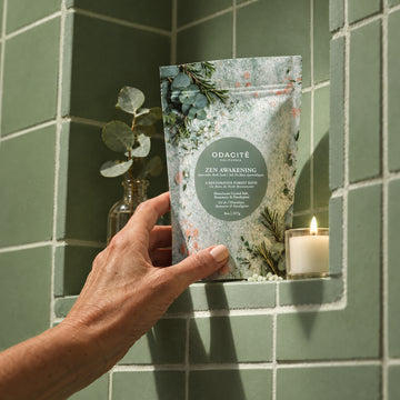 A hand holds a Zen Awakening • Himalayan Crystal Salt, Rosemary & Eucalyptus • Ayurvedic Bath Soak pouch in front of a green wall and vase.