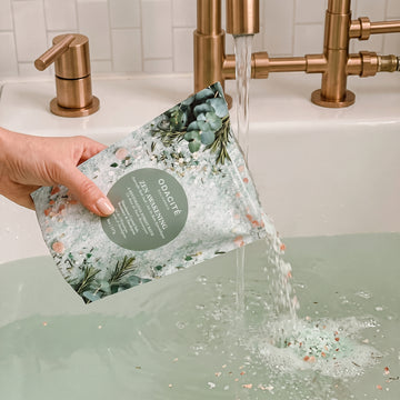 A hand pours Zen Awakening • Himalayan Crystal Salt, Rosemary & Eucalyptus • Ayurvedic Bath Soak from a pouch into a bathtub faucet.