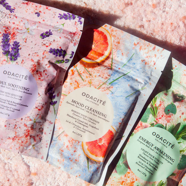 Three Odacité Relax & Recenter Bath Set packets with floral and fruit patterns on a pink background.