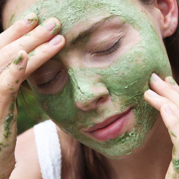 Woman in a white top applies a green face mask with eyes closed after using Odacité Uneven Skin Double Cleansing Set.