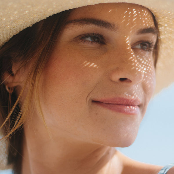 Woman in sunlight with glowing skin, wearing a straw hat and holding Odacite Skin Glow Serum Set.