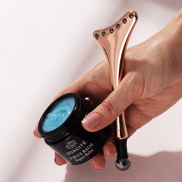 Hand holds Odacité CALM & COOL CRYOTHERAPY DUO next to an open jar of blue repair balm on a light background.
